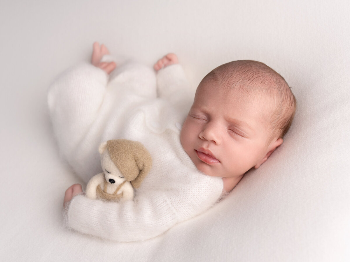 Nyfødt baby i hvit heldress sover fredelig med en liten bamse under nyfødtfotografering nær Oslo.
