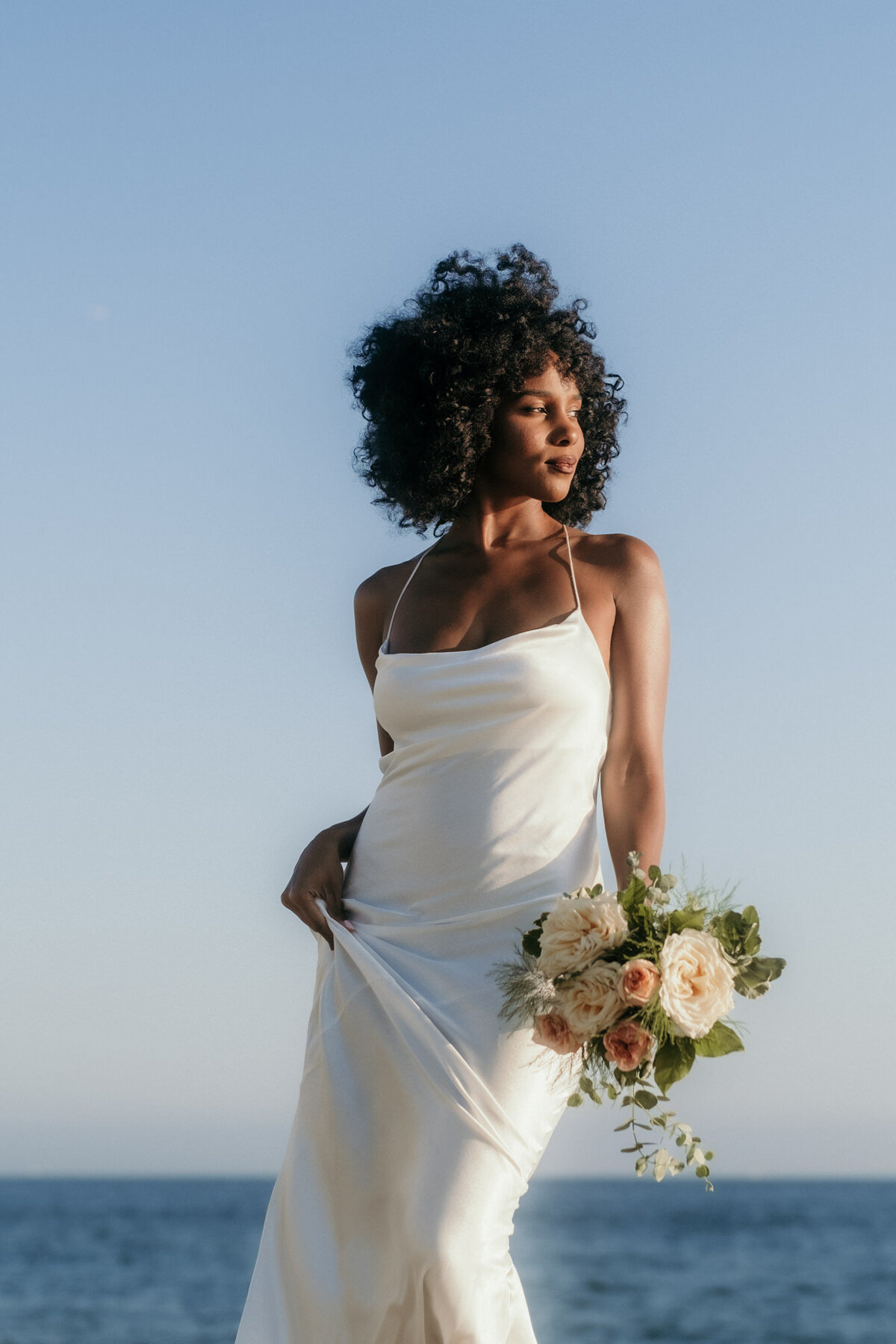 Mit dem Meer im Hintergrund posiert die Braut mit ihrem Strauß in einer Hand.