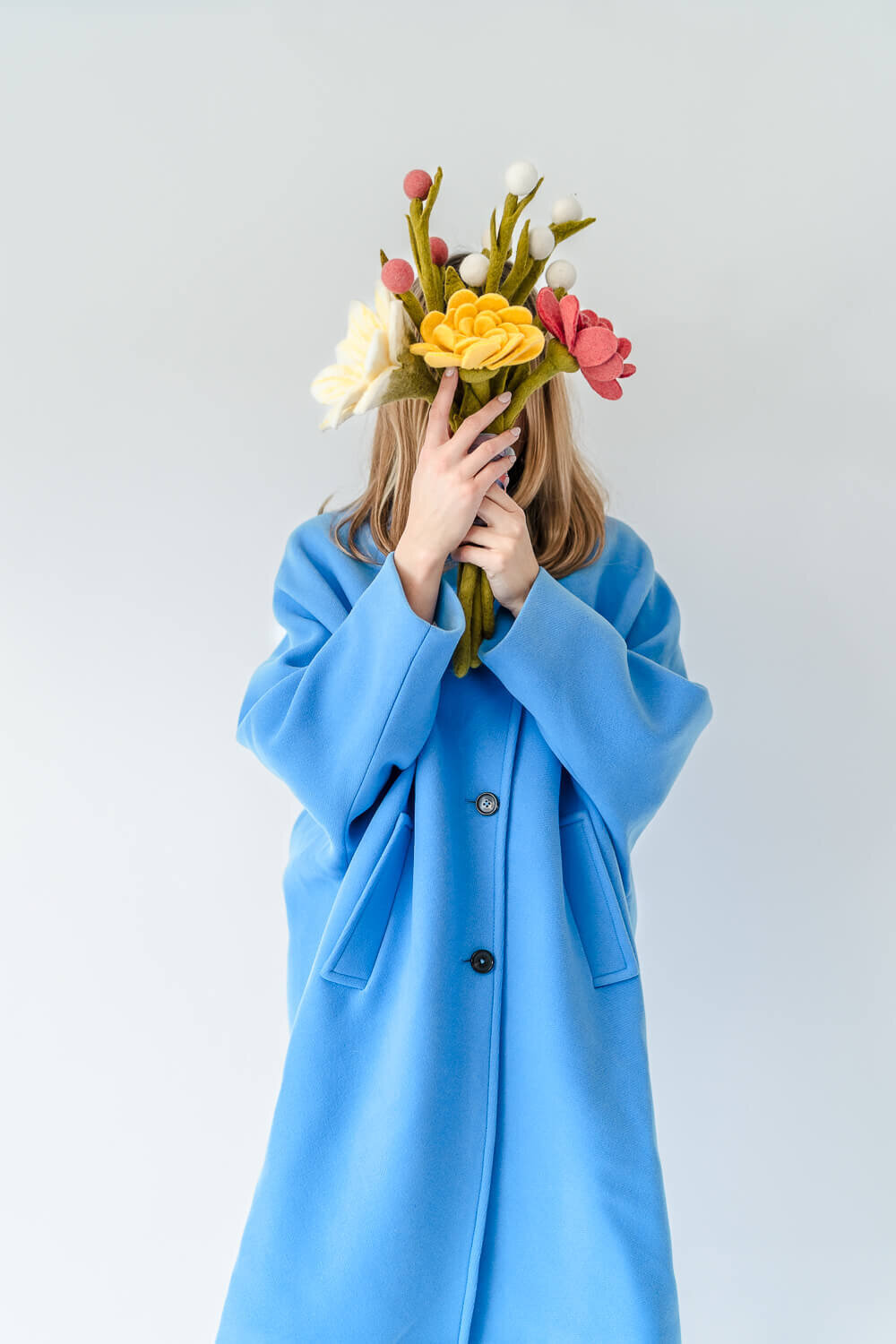 Woman wearing bright blue coat holding large bunch of colourful felt flowers in front of face.