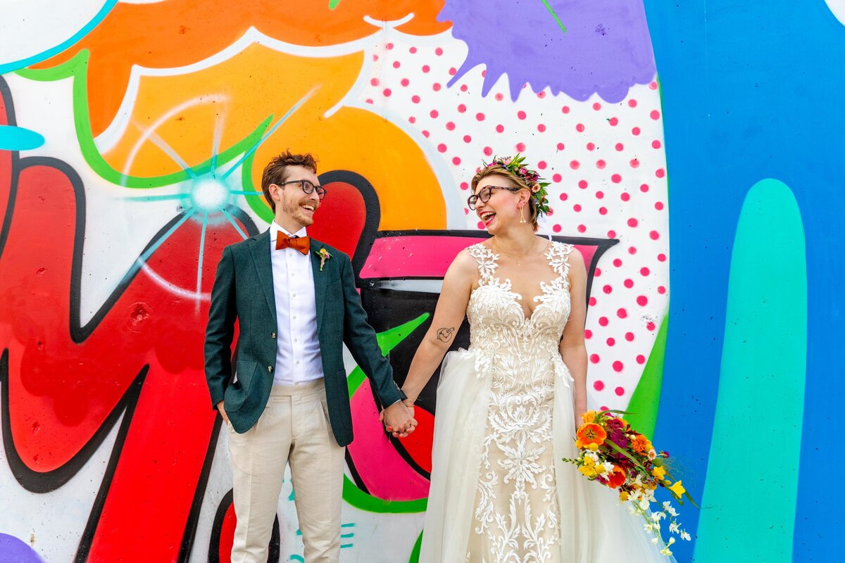 A wedding couple holding hands and standing in front of a colorful mural.