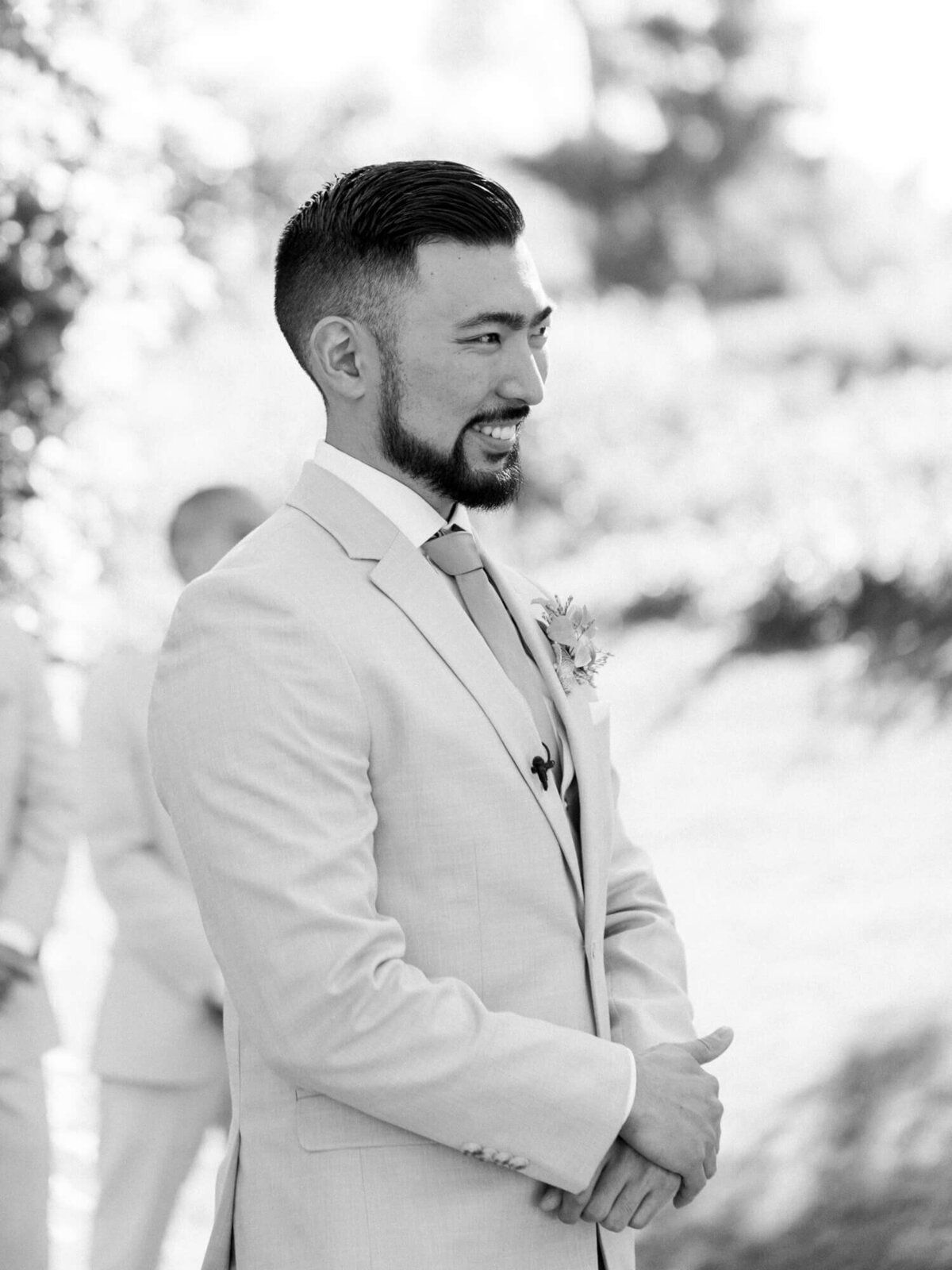 A groom in a light suit smiles warmly, standing outdoors in a sunlit garden. The background is blurred, highlighting his joyful expression.
