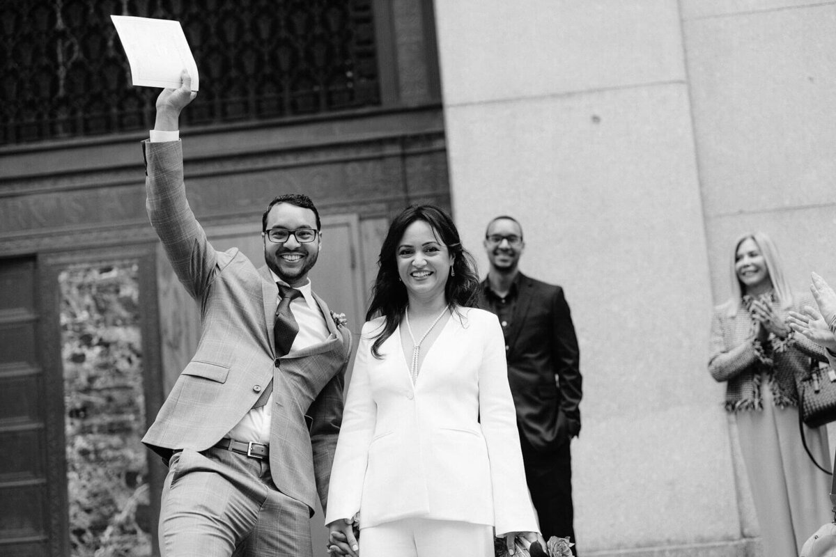 The groom is excitedly waving their marriage certificate in the air, as he and his bride exit NY City Hall. Image by Jenny Fu Studio