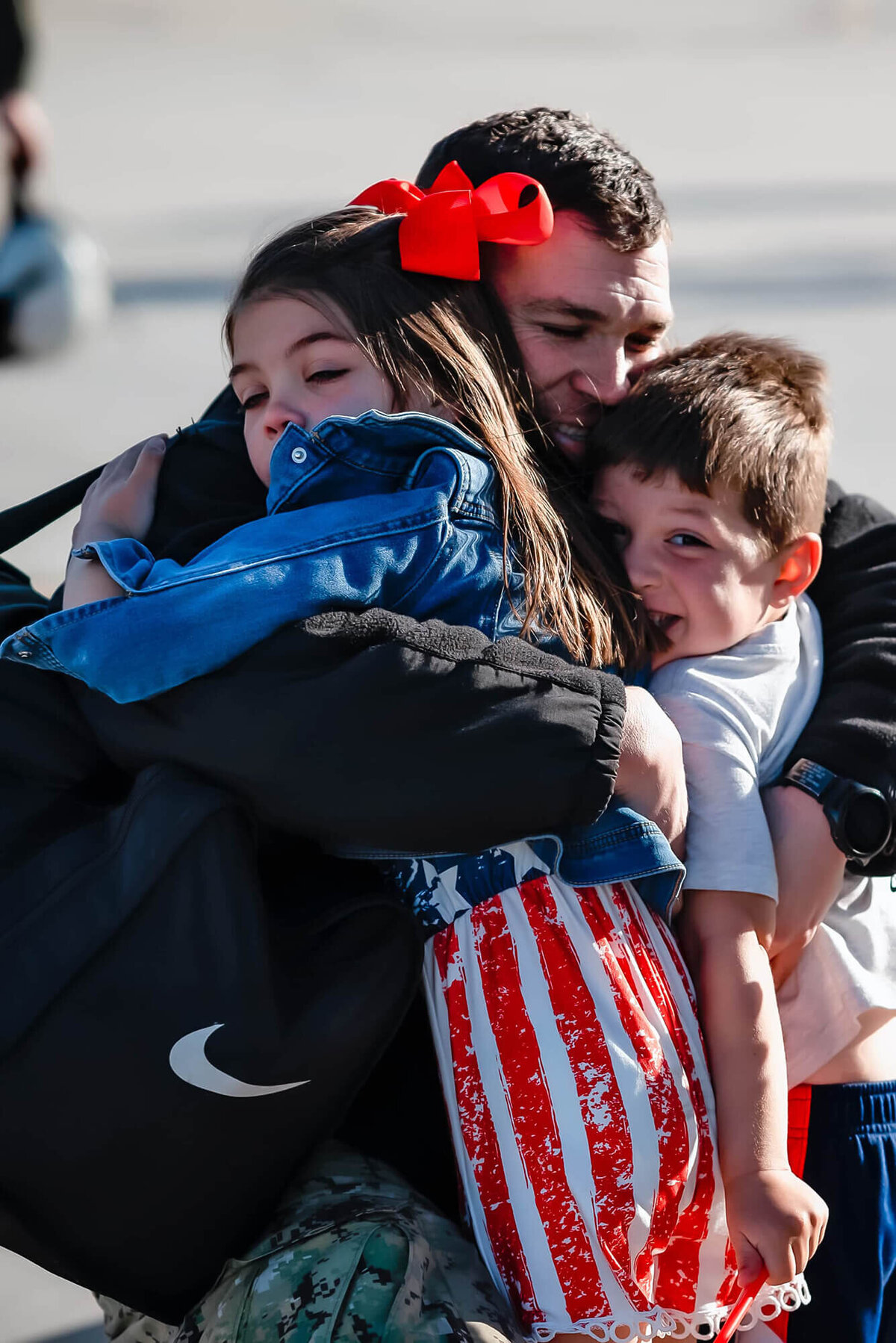 Two young kids, wearing red, white, and blue give their dad a big hug as they welcome him home from deployment.