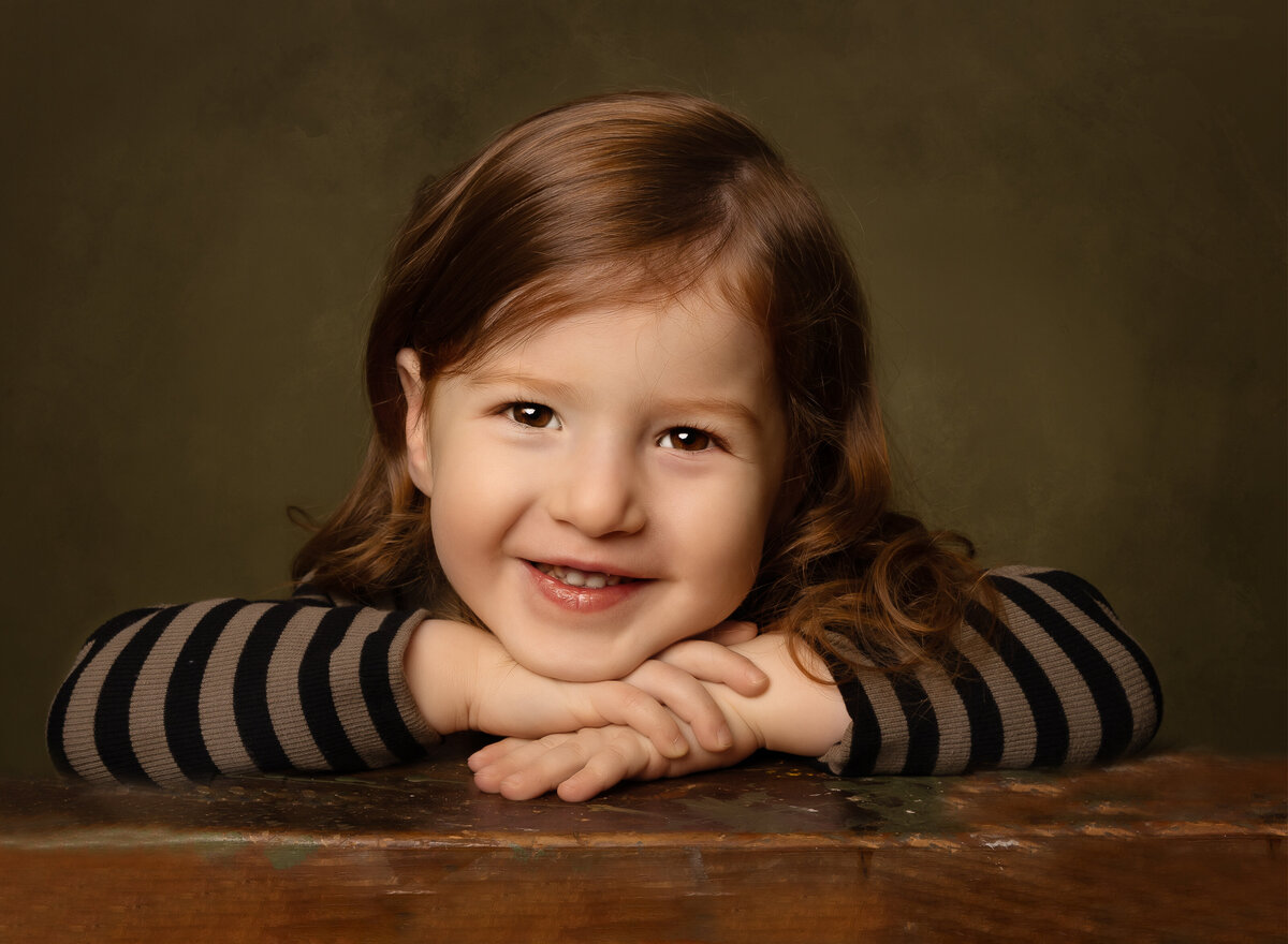 Little boy is posing for a studio portrait in brooklyn to commemorate upsherin. His hands are folded under his chin and he is smiling at the camera. Captured by brooklyn family photographer Chaya Bornstein.