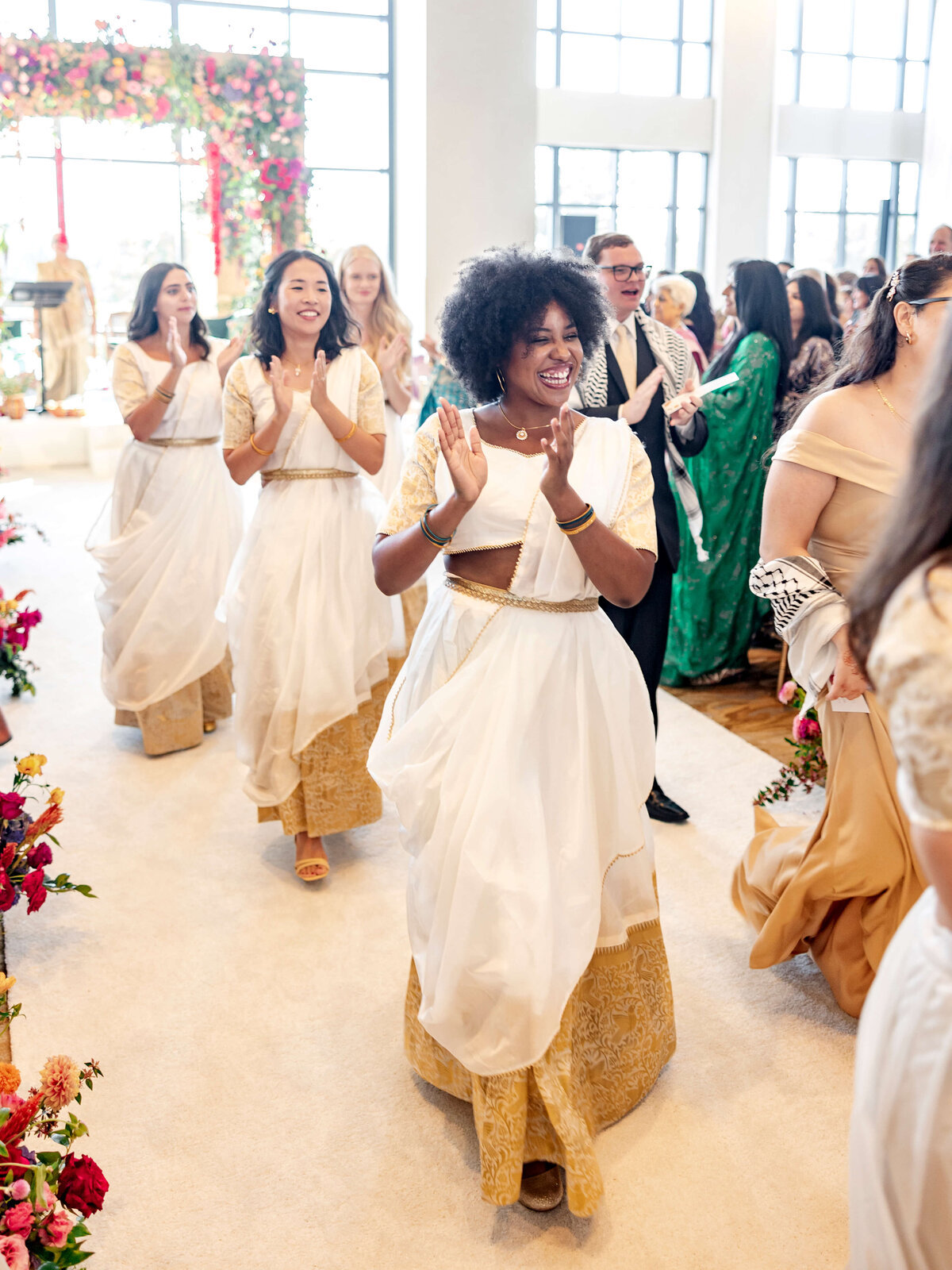 A joyful procession of people smiling and clapping, wearing elegant white and gold attire, walk down an aisle adorned with colorful flowers. Bright light streams in from large windows, creating a celebratory atmosphere.