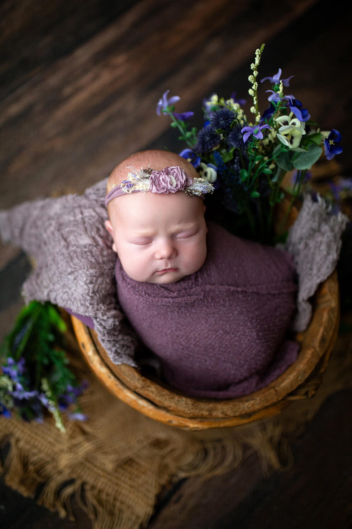 Newborn baby wrapped in purple in brown bucket surrounded by purple flowers