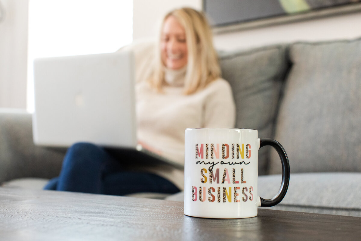 A close-up of a white mug that reads “Minding my own small business” with a blurred female entrepreneur working on her laptop in the background—capturing the essence of small business life.
