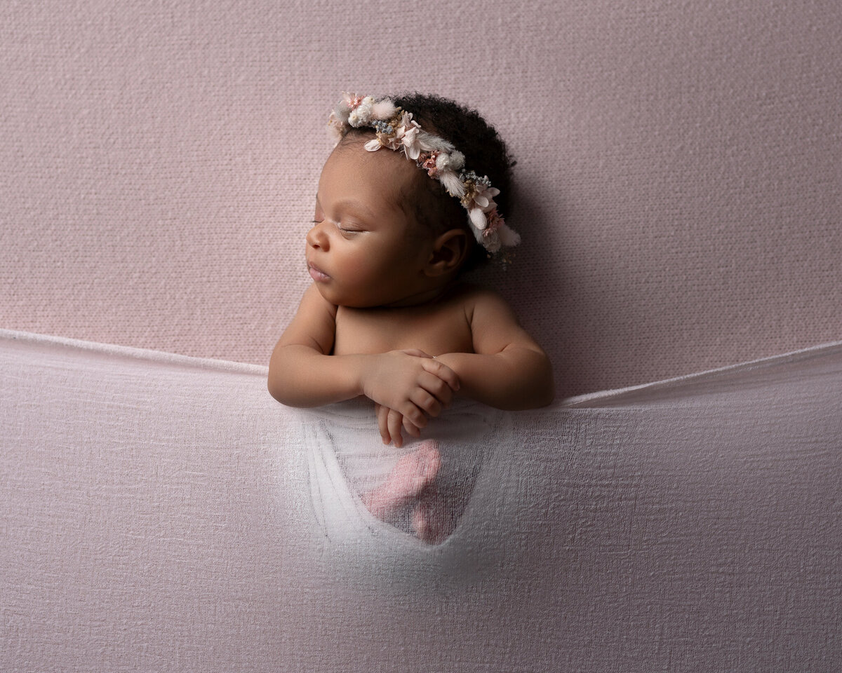 Baby girl posed with hands under chin and floral crown on soft purple backdrop – newborn photo studio in Austin TX