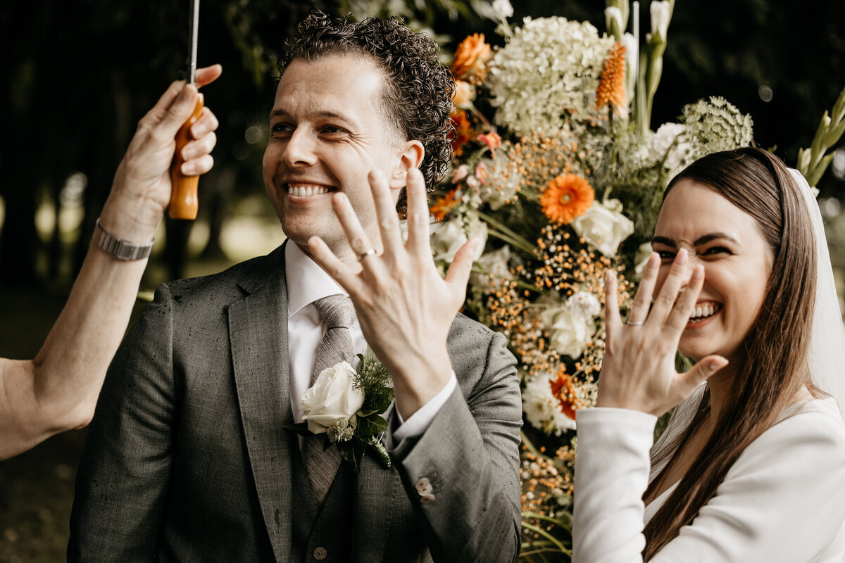 Het bruidspaar straalt terwijl ze trots hun trouwringen laten zien, omringd door kleurrijke bloemen.