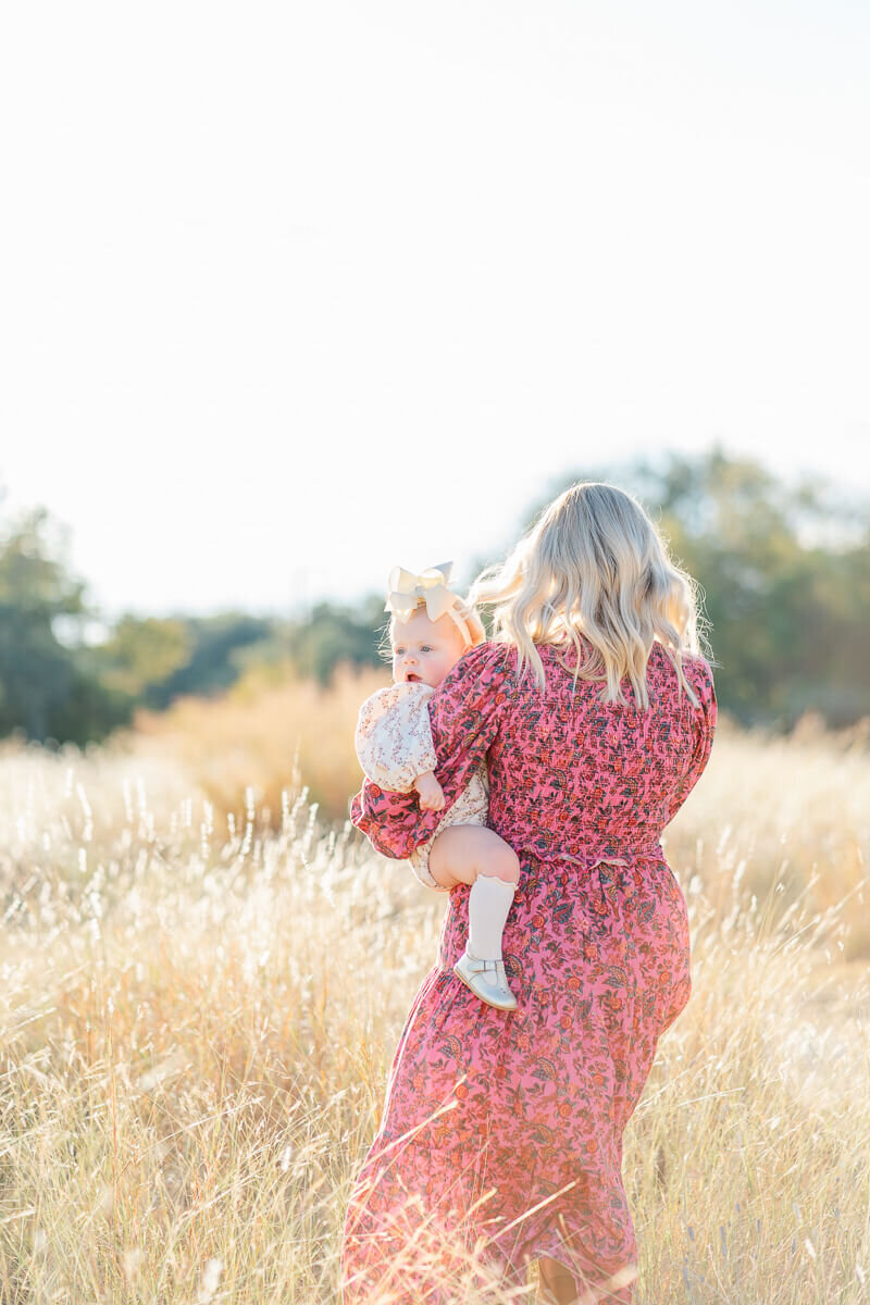 a mother in a long burgundy dress holds her daughter and walks through a field of golden grass during her family session in Austin. 