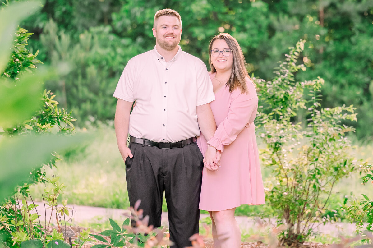 A happy couple holding hands underneath greenery at Daniel Stowe Botanical Gardens in the springtime during their engagement session by JoLynn Photography, a Raleigh wedding photographer