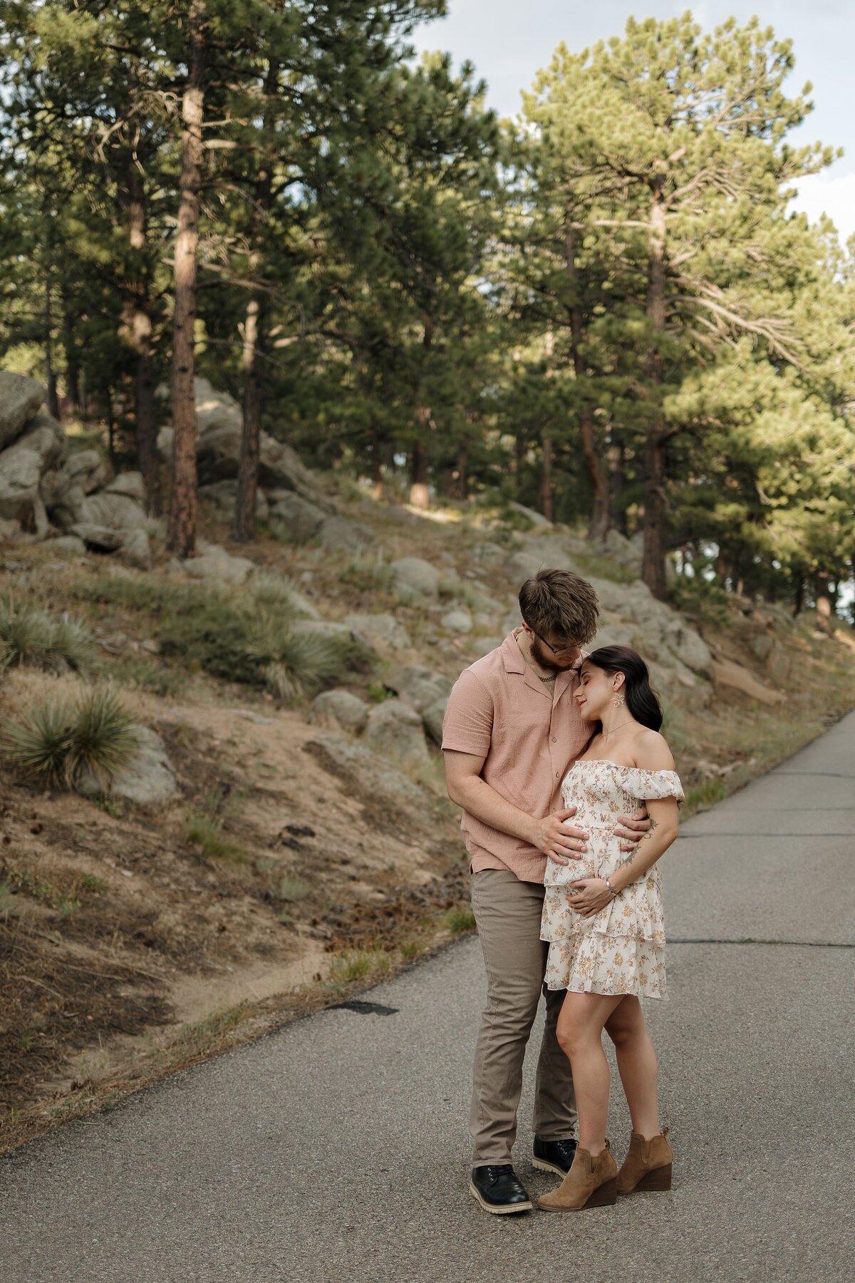expectant mom resting head on dads shoulder while dad has hand on belly in front of a row of trees in boulder colorado