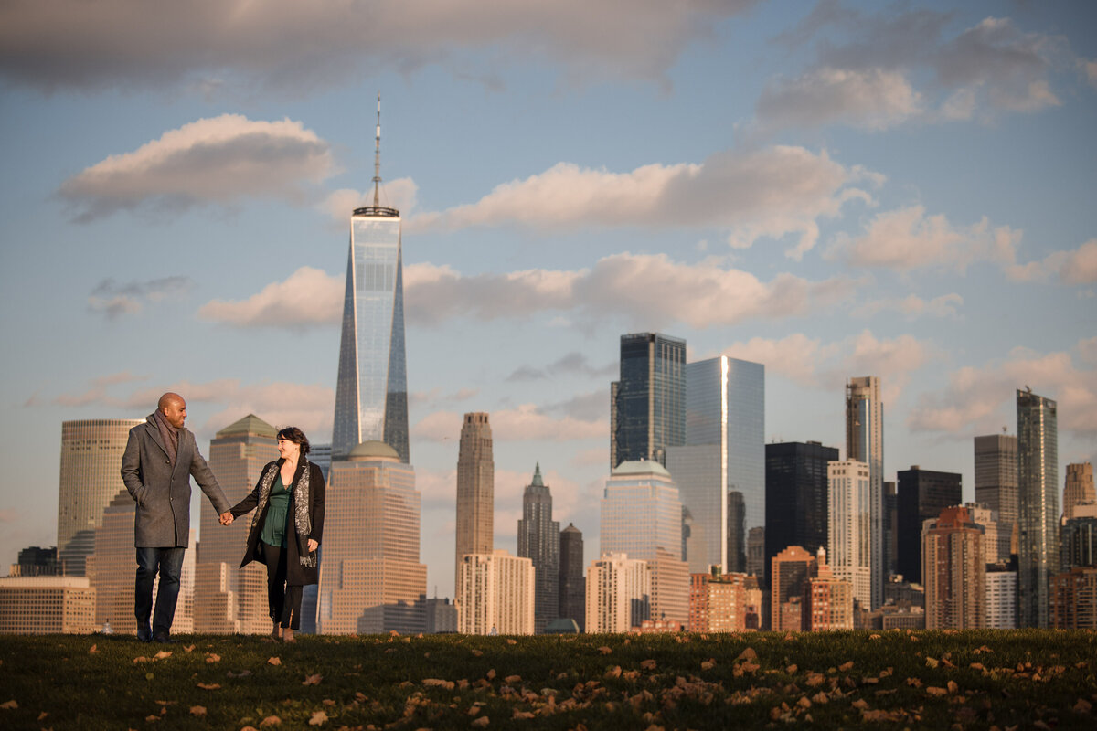 A couple holding hands and walking in a park with a view of the New York City skyline behind.
