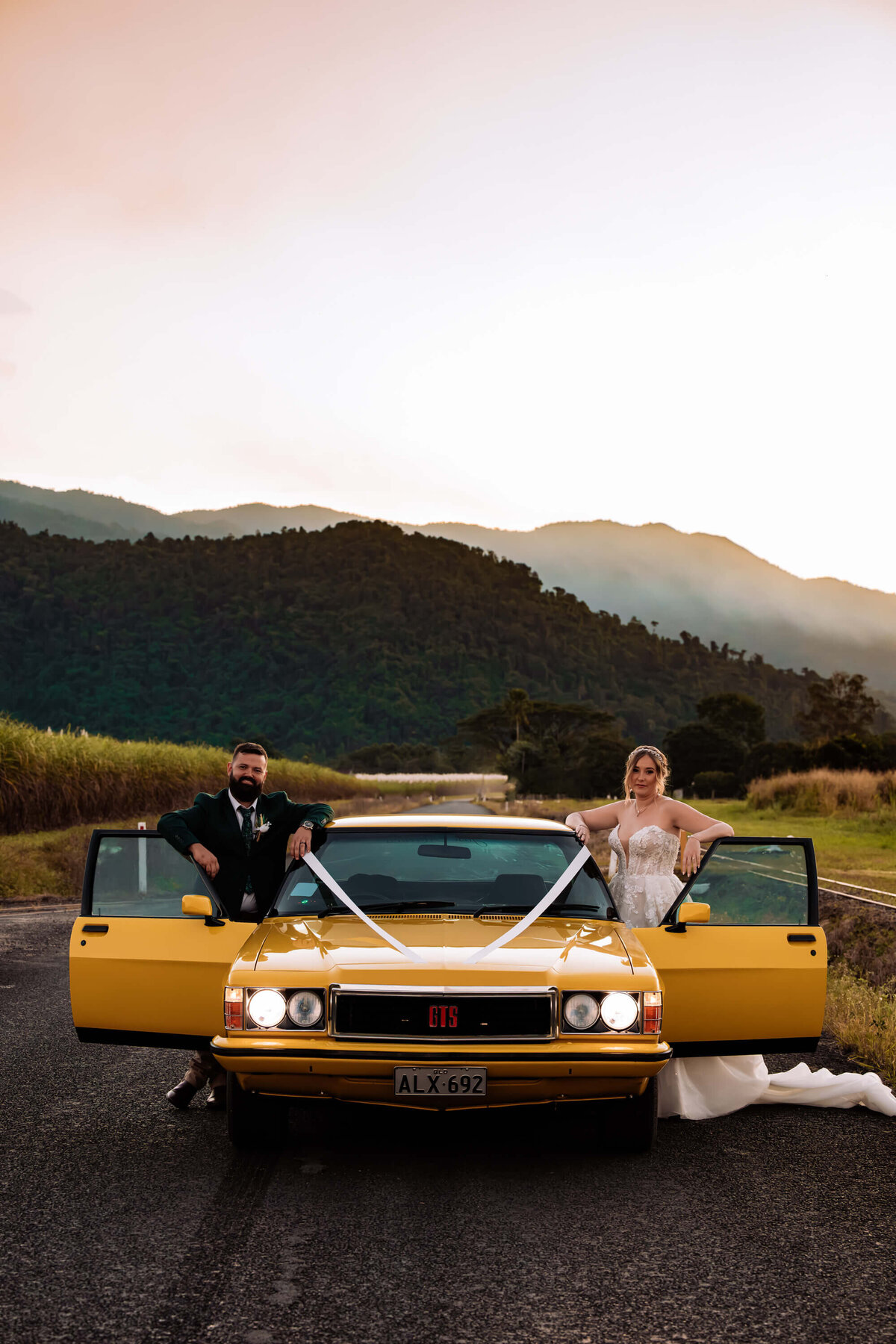A joyful bride and groom pose beside their bright yellow car on a scenic road, capturing a moment of love and celebration. - photo by Jamie Simmons
