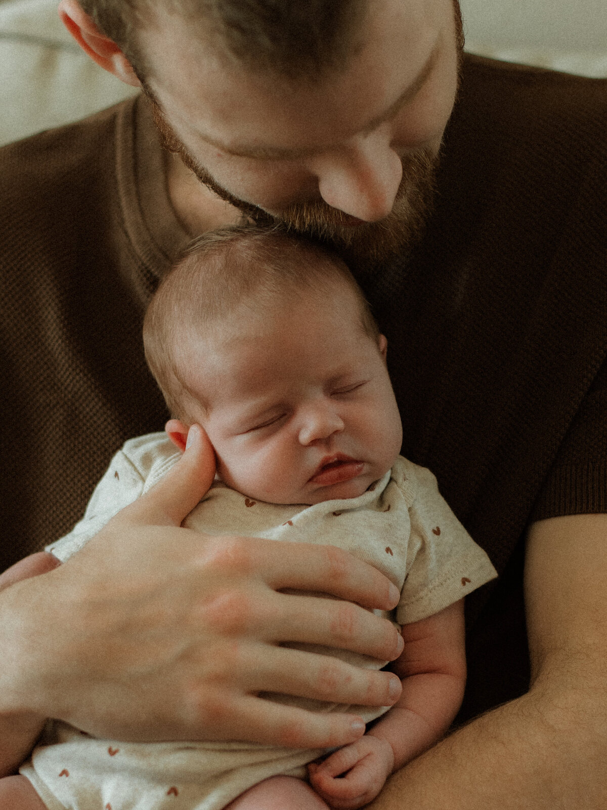Father lovingly cradling his sleeping newborn in his arms, resting his cheek against the baby's soft head in a serene and intimate scene.