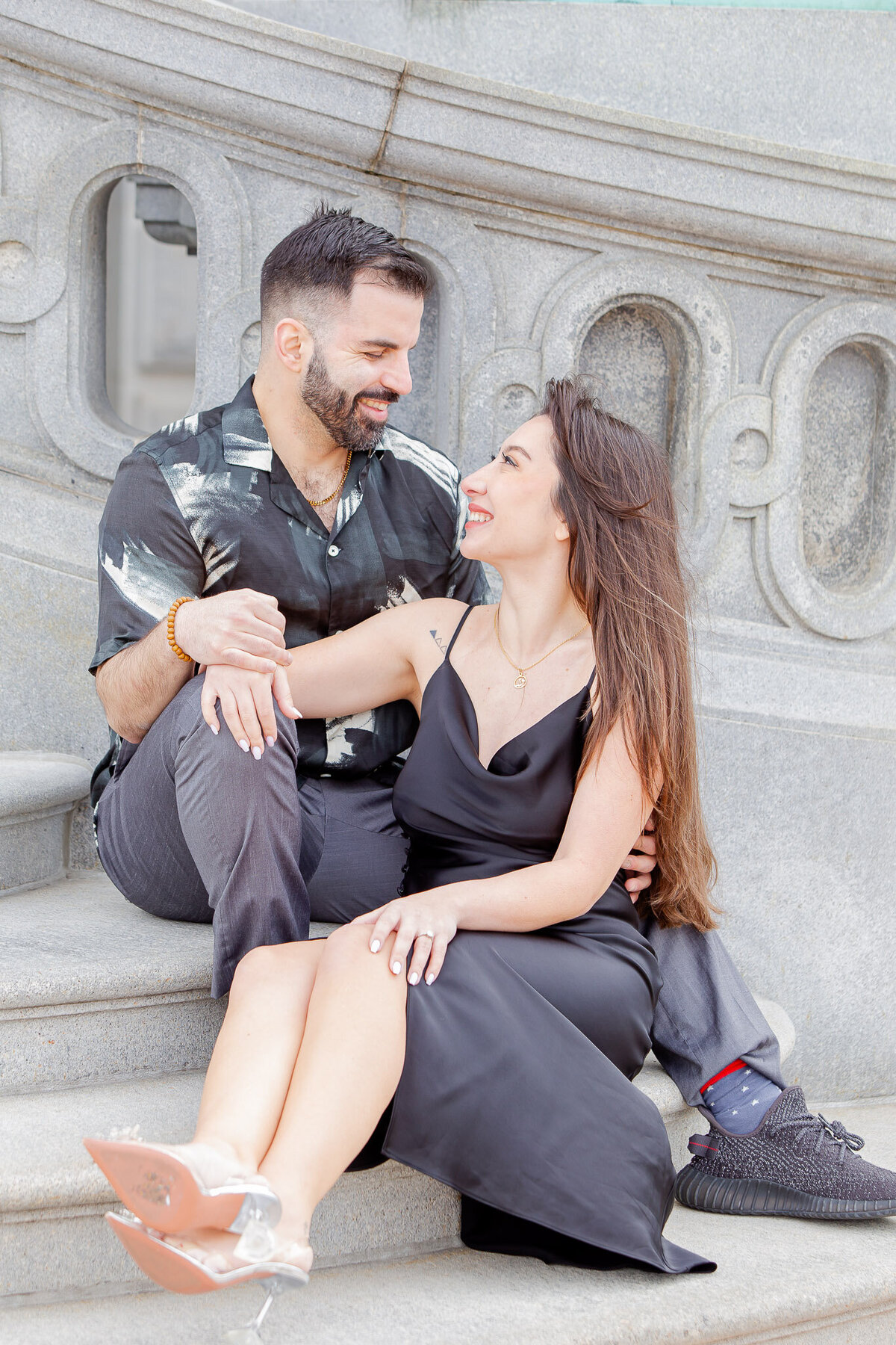 A man and woman sit on stairs outside in downtown Raleigh. She is wearing a simple black dress and he is also wearing all black.