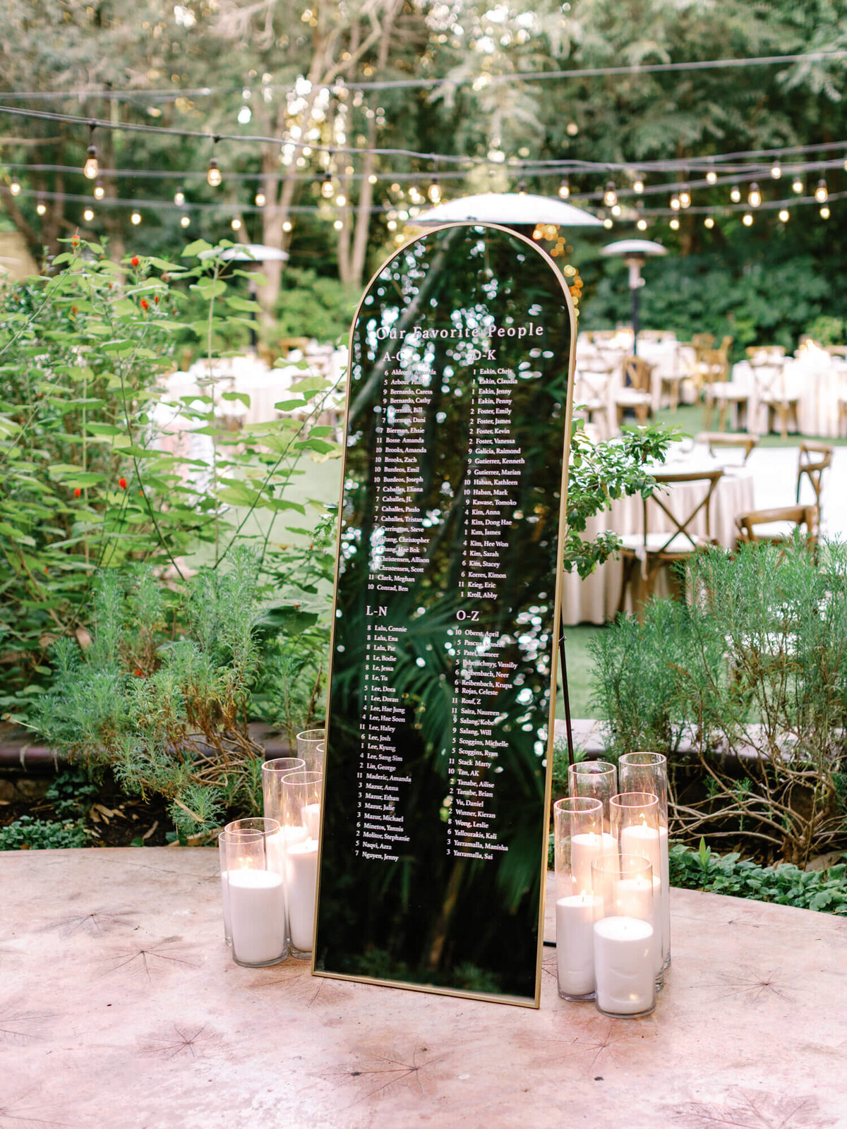 Tall black seating chart with white text listing guest names, surrounded by lit candles on a wooden table.