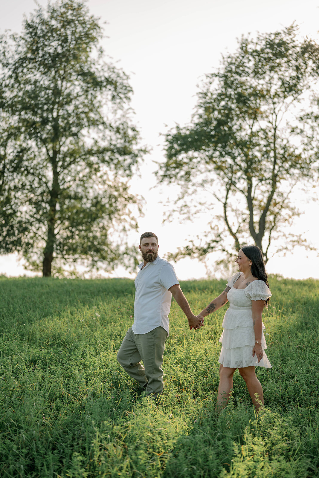 Kali and Joe holding hands while walking through a green field during their summer engagement session near Detroit.