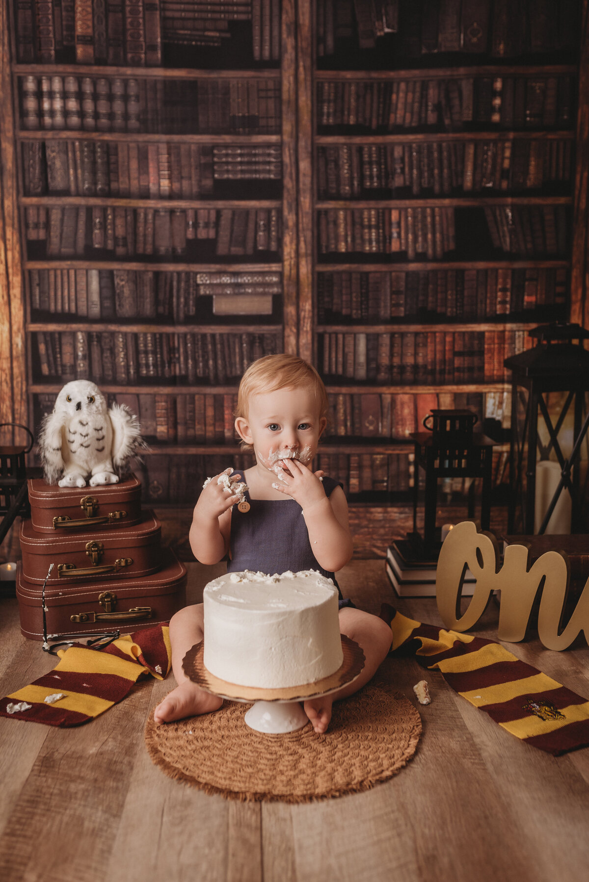 Harry Potter themed cake smash portrait session book case in background with stack of suitcases and white snowy owl on top, Happy Potter scarf around baby with the letters O N E and one year old boy eating white cake with his hands licking his fingers