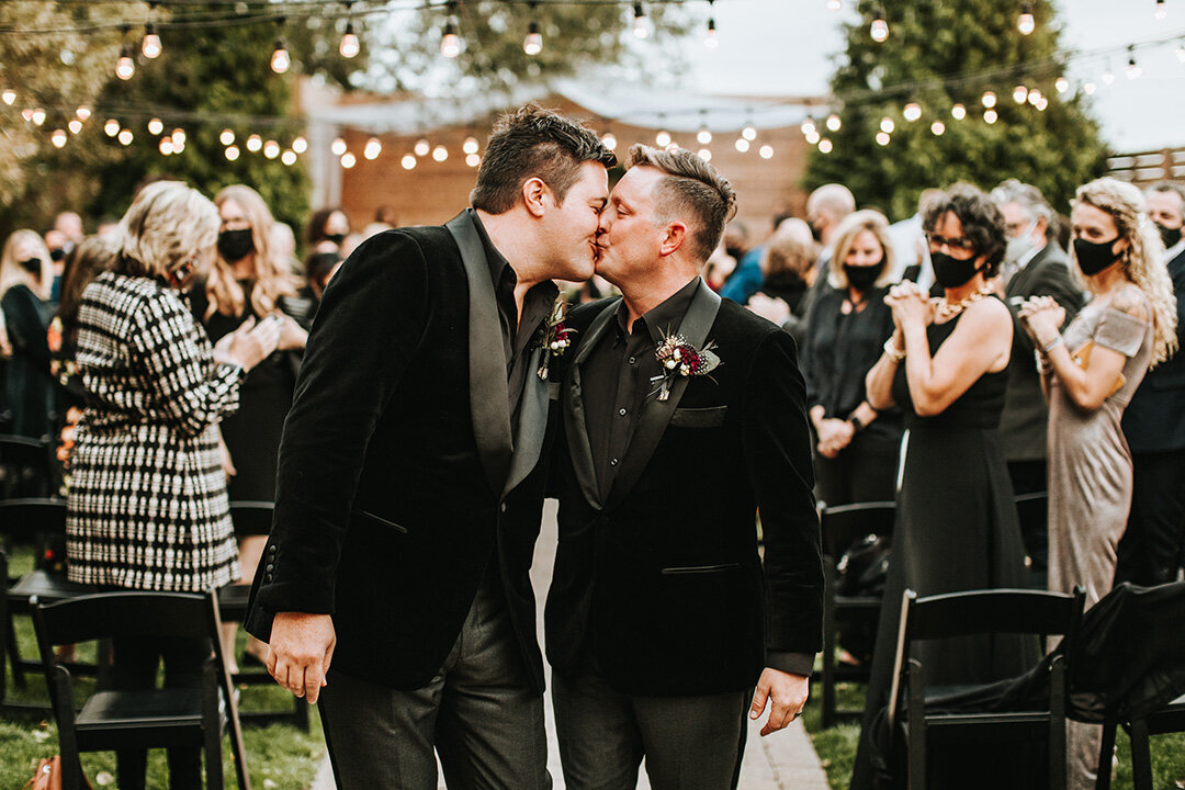 Two grooms wearing black tuxedos share a kiss at their wedding while guests look on in the background.