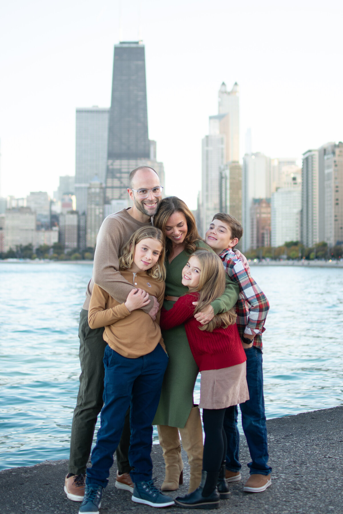 candid family photo of parents and three children embracing by Lake Michigan, with the Chicago skyline in the background, taken during golden hour family photography session