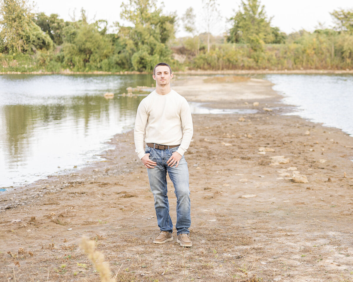 A high school senior male, standing next to a pond, serious look at the camera, with short brown hair, wearing a light tan long sleeve shirt. blue jeans, light brown dress shoes, a belt.