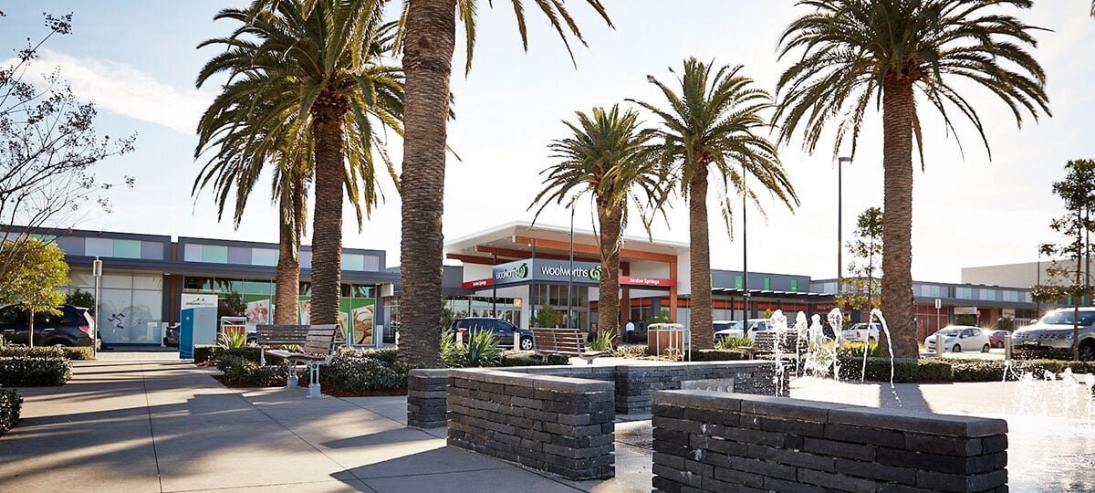 Wide street-level image of Jordan Springs Shopping Centre featuring Woolworths entryway, tall palm trees, and surrounding landscape elements. Built by Serlana Construction.