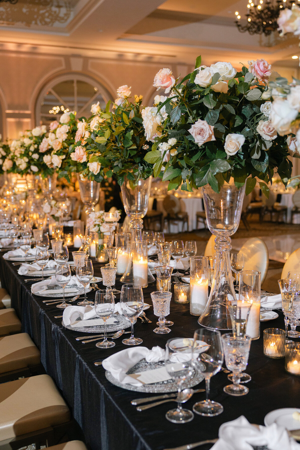 elegantly styled wedding reception table at The Adolphus in Dallas featuring a black tablecloth, tall glass vases with pink rose arrangements, numerous candles, and refined glassware and chargers