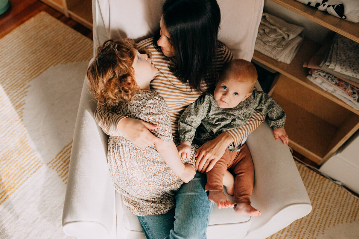 A mother snuggles her two young daughters in a cream colored rocker in their nursery