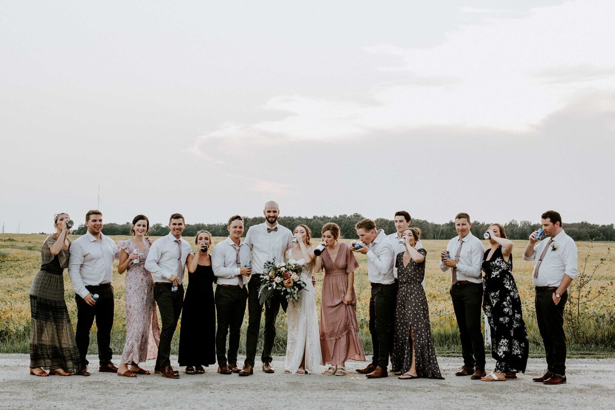 Bride, groom, and wedding party are standing in a line drinking a beer for a fun portrait at a rustic farm wedding in Exeter, Ontario