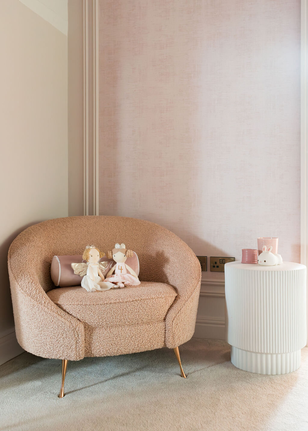 A cozy pink chair in the corner of a girl's soft pink bedroom. The walls are wallpapered in light pink, and there is a modern white side table next to the chair.