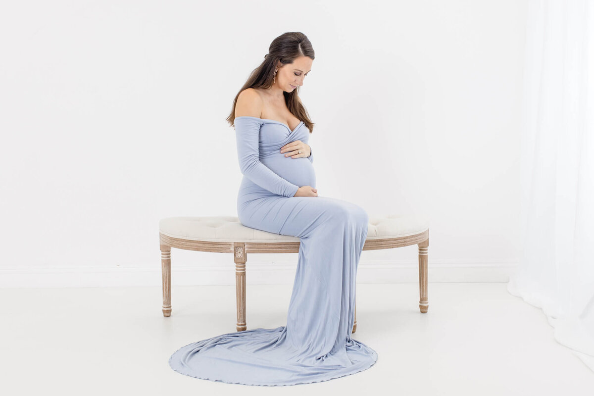 A pregnant woman in a long, light blue dress sits on a bench in a bright, minimalist room. She holds her belly with both hands and looks down, surrounded by soft, white light and decor.