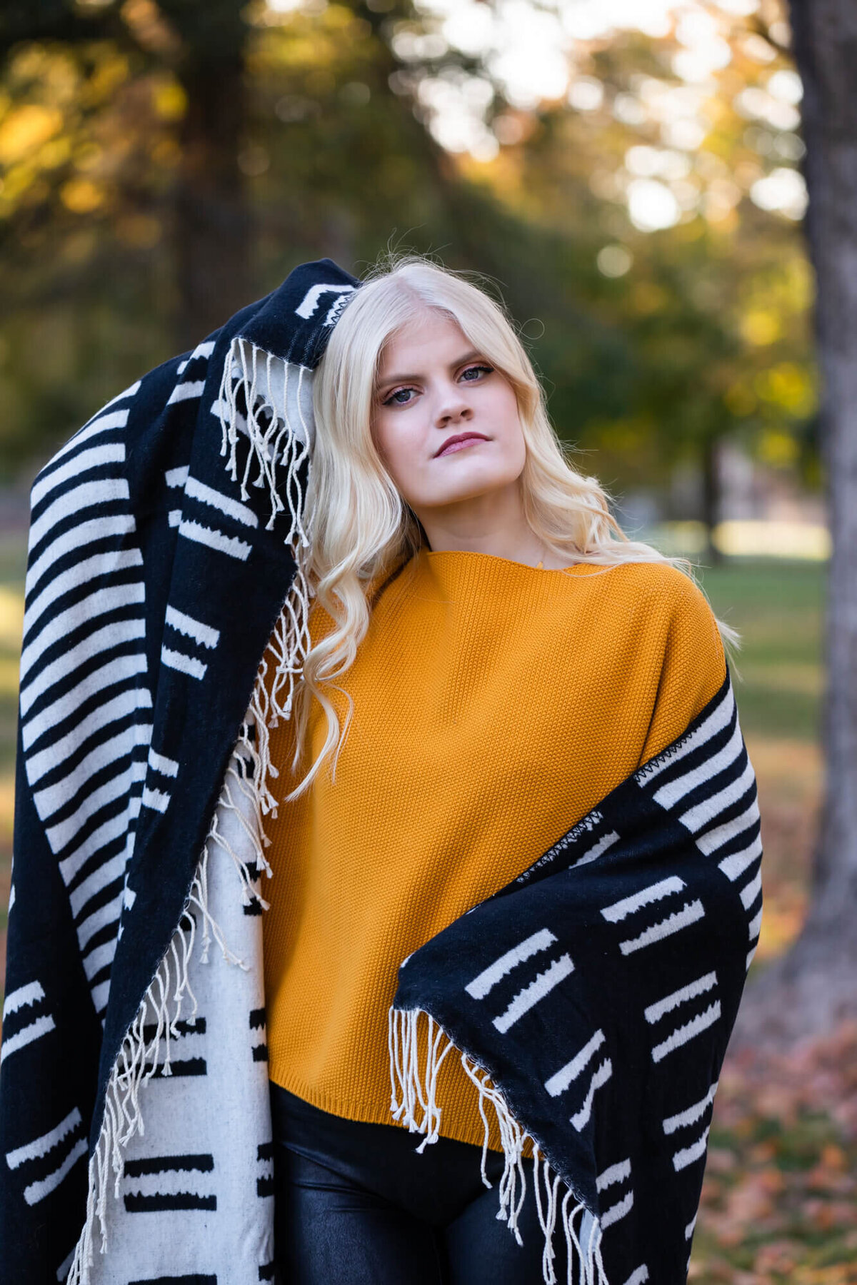 A Fall senior picture of a beautiful blonde teen girl wearing a dark yellow sweater and wrapping herself in a black and white blanket. Captured by Springfield, MO senior photographer Dynae Levingston.