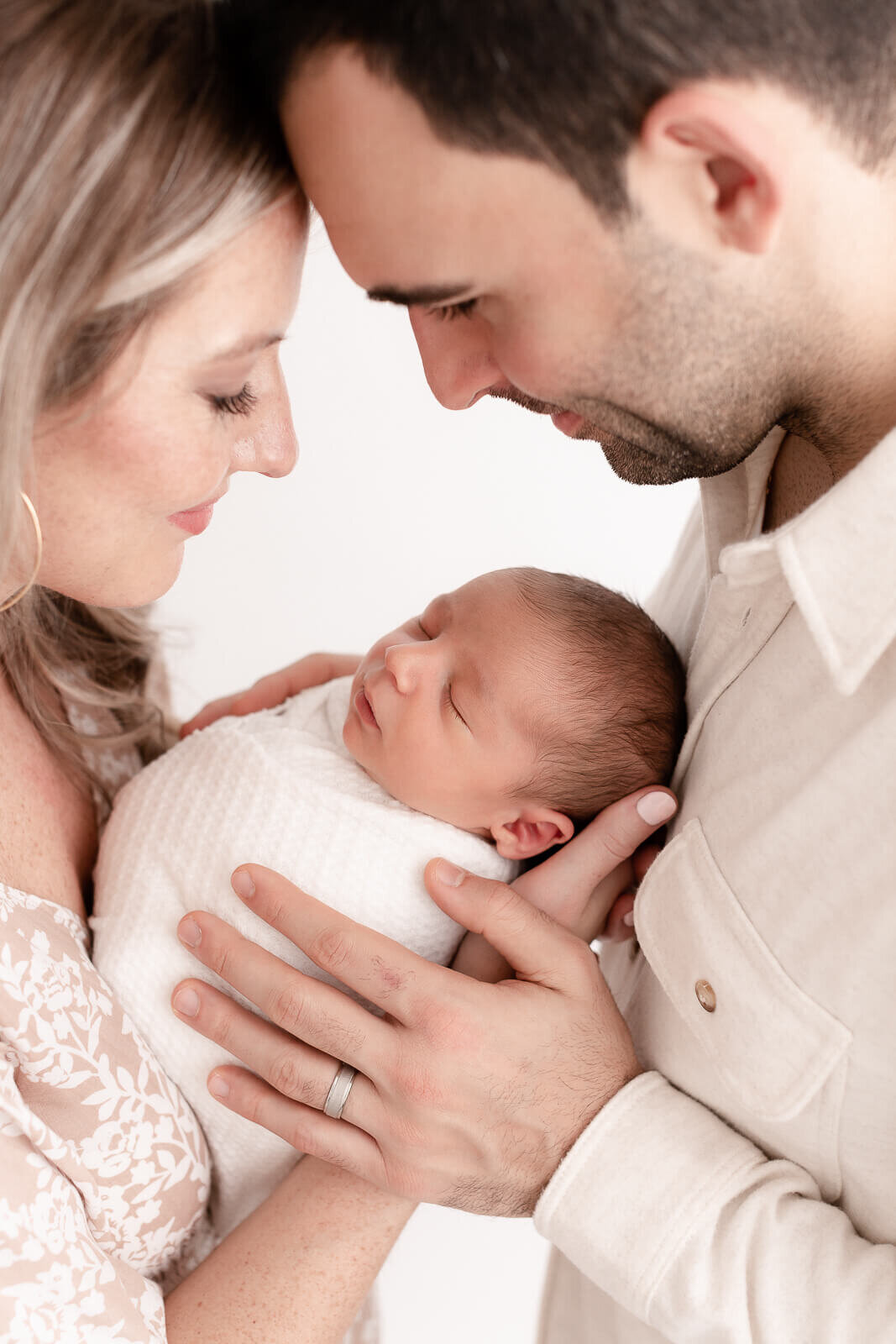 New Mom and Dad standing face to face and touching foreheads while holding their baby in between them. They are both smiling down at their baby while baby sleeps peacefully.