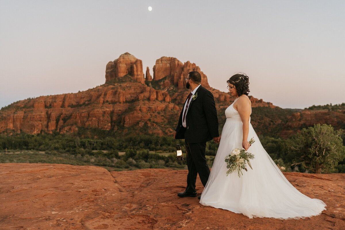 A newlywed couple holding hands and walking in front of red rocks 