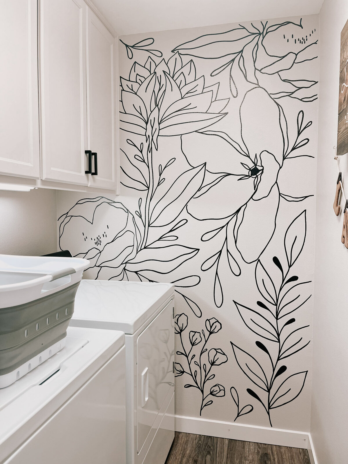 A laundry room with a basket and two machines featuring a wall in the back that has custom florals painted on it representing full service design and calligraphy by Life in Letters.