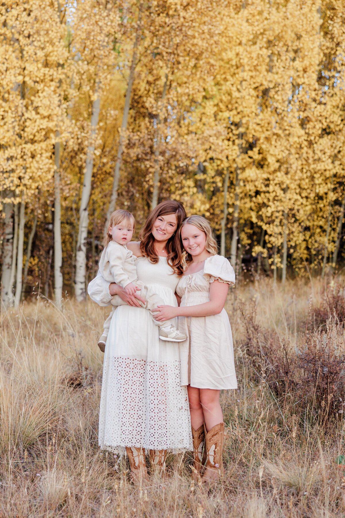 A smiling mom hugs her baby on her hip and teen daughter in an Aspen forest in fall