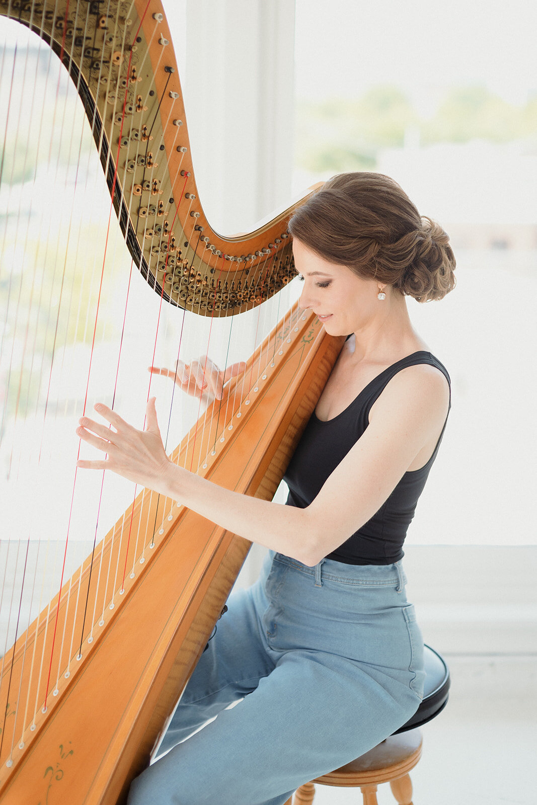 fair skinned woman with dark hair in black tank top and jeans, playing harp.