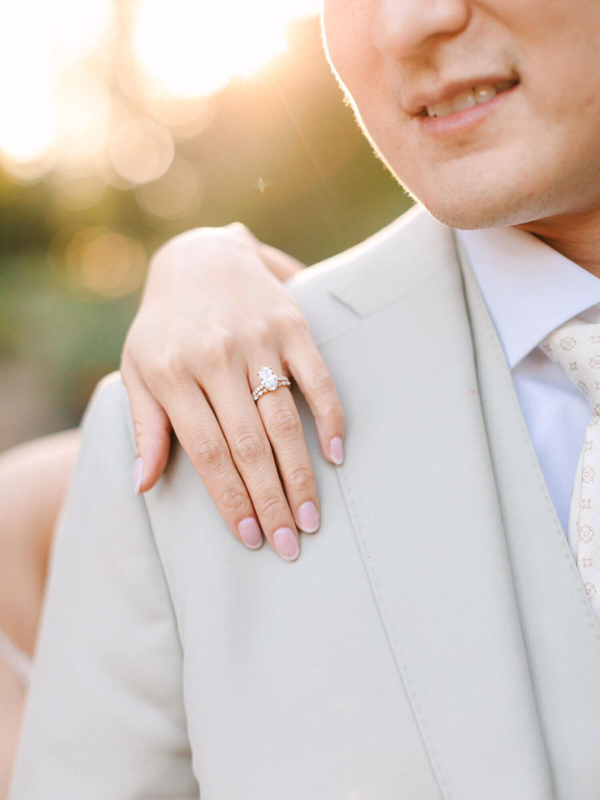 Close-up of a hand with an engagement ring on a suited shoulder.