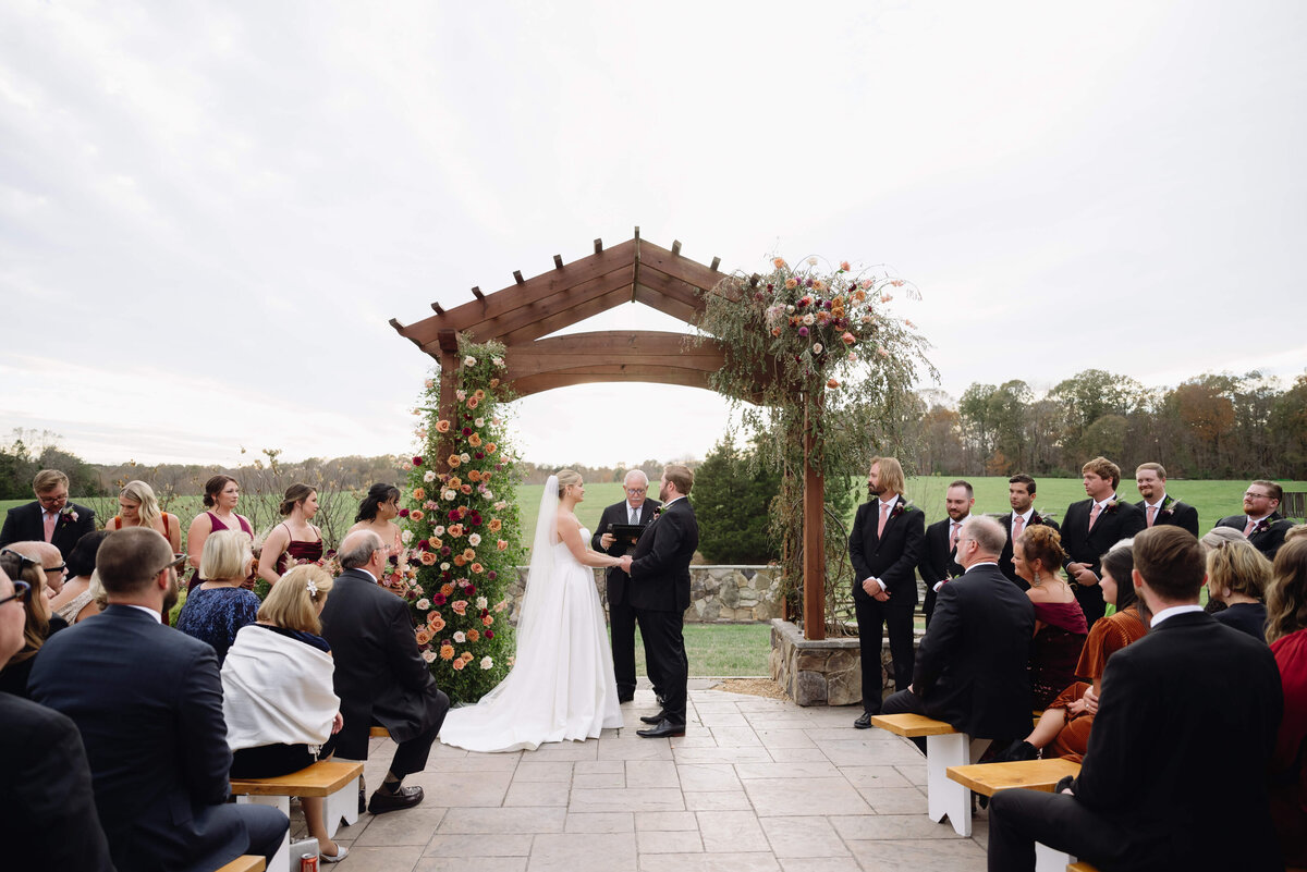outdoor wedding ceremony with bride and groom holding hands under a wedding arch decorated with pink and orange florals at Layz S Ranch with the sun setting in the distance over a tree line photographed by Virginia wedding photographer