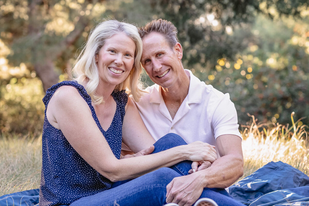 A husband and wife with blonde hair sitting on a navy picnic blanket in green grass wearing shades of blue and white looking at the camera with their heads together at Jeffrey Open Space in Irvine.