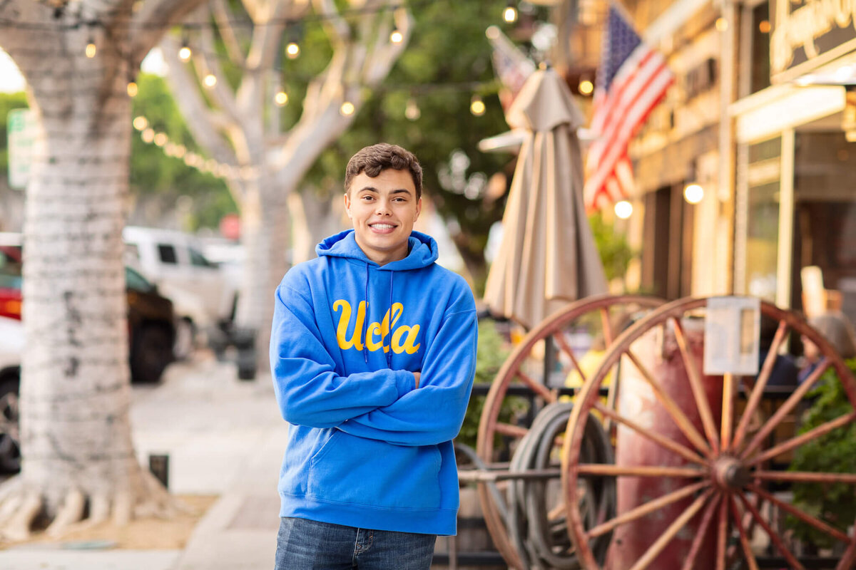 A young person in a blue UCLA hoodie stands with arms crossed on a quaint street lined with string lights. An American flag and rustic wagon wheel are visible in the background.