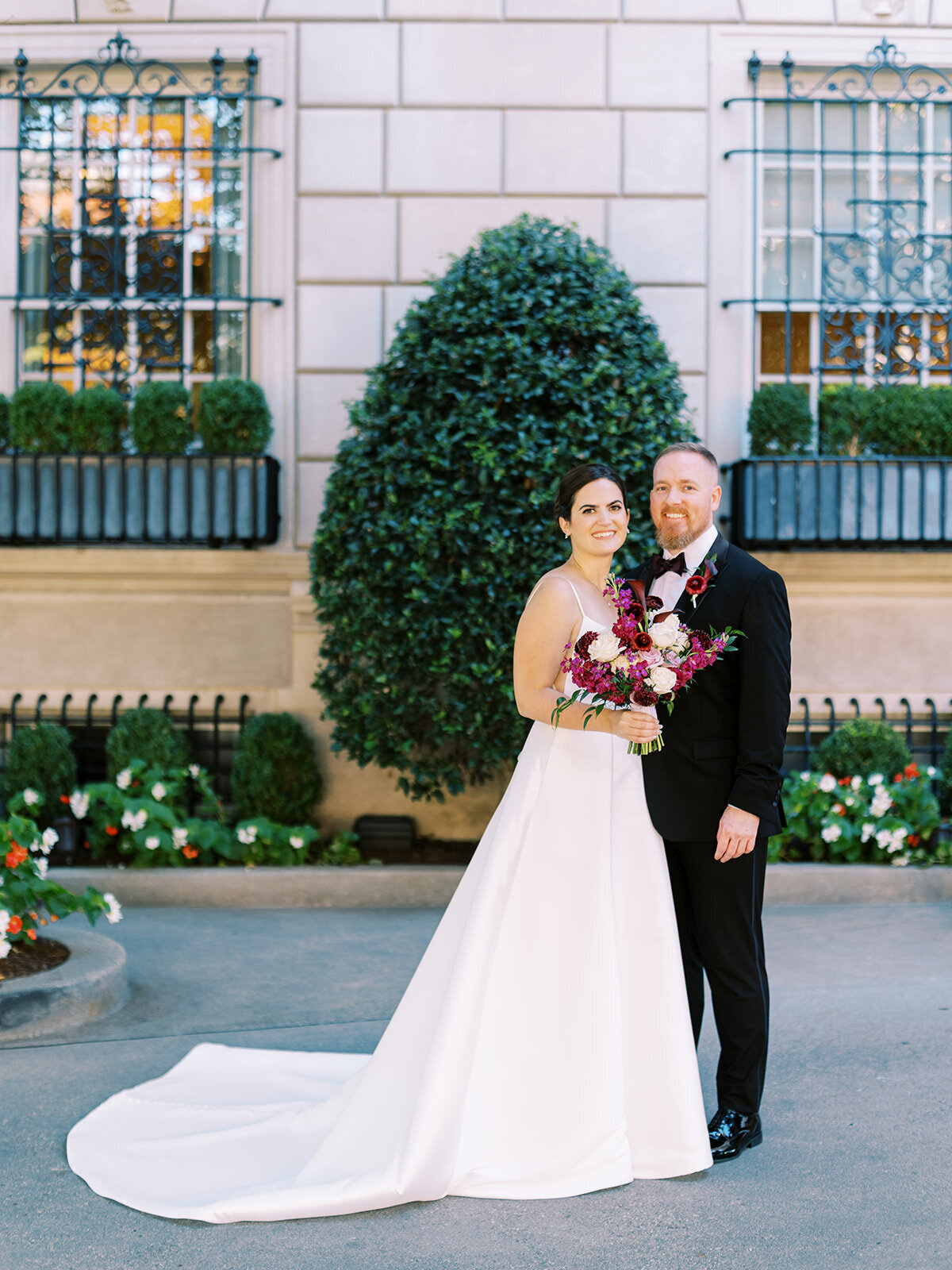 Bride and groom standing outside an elegant Hay Adams DC, with the bride holding a vibrant bouquet of pink, burgundy, and purple flowers