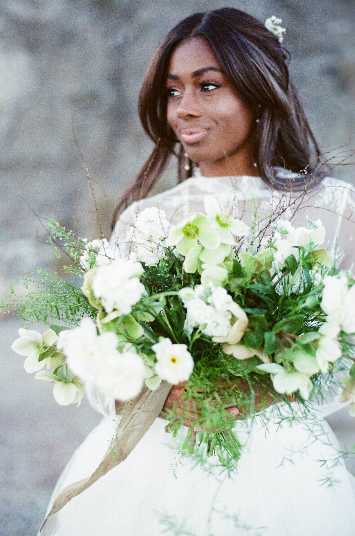 A bride with dark skin, wearing a white wedding gown and holding a bouquet of white flowers, gazes into the distance during her portrait session.