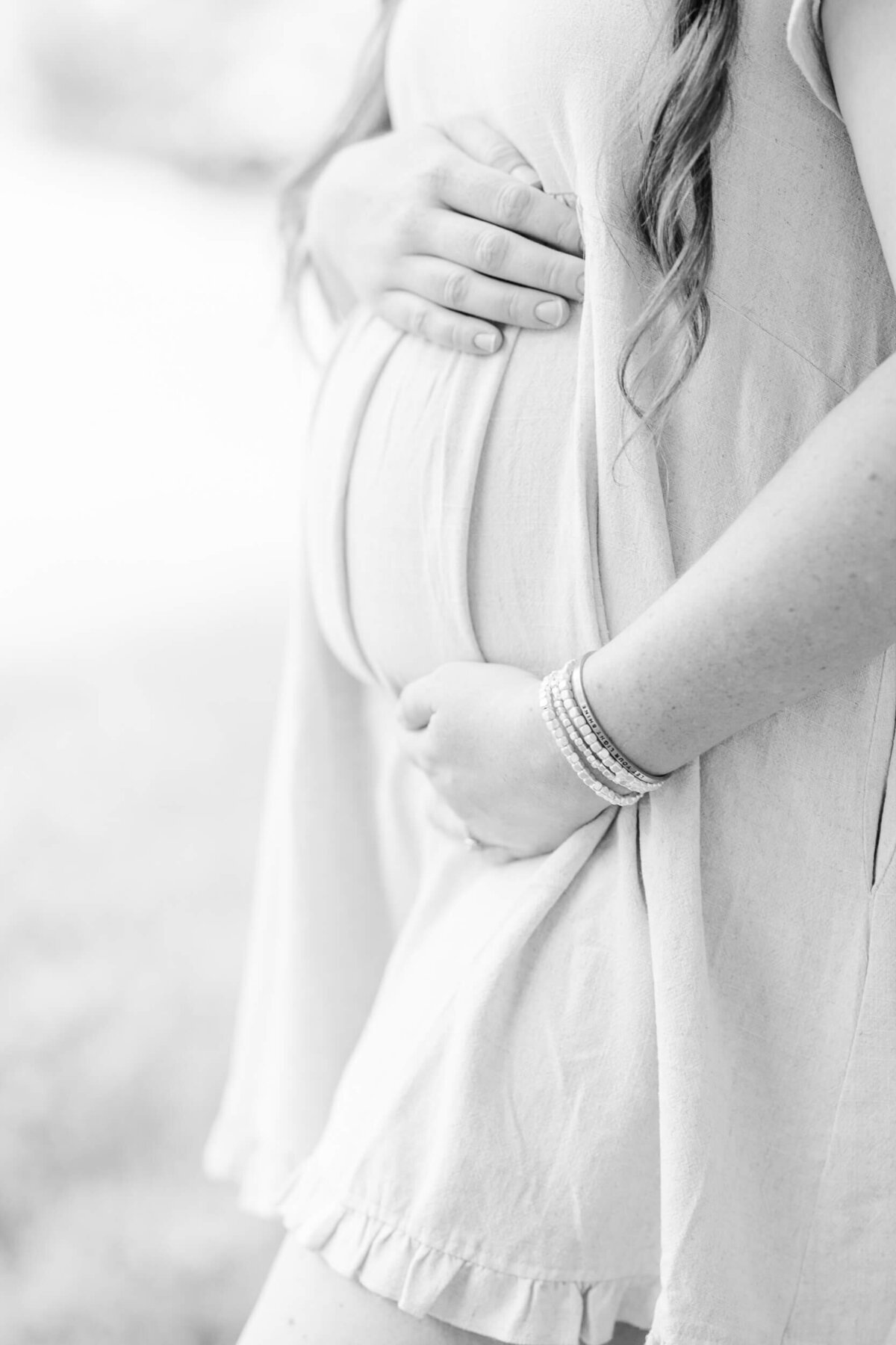Black and white, close up of a woman cradling her baby bump with a bracelet that says "let your light shine"