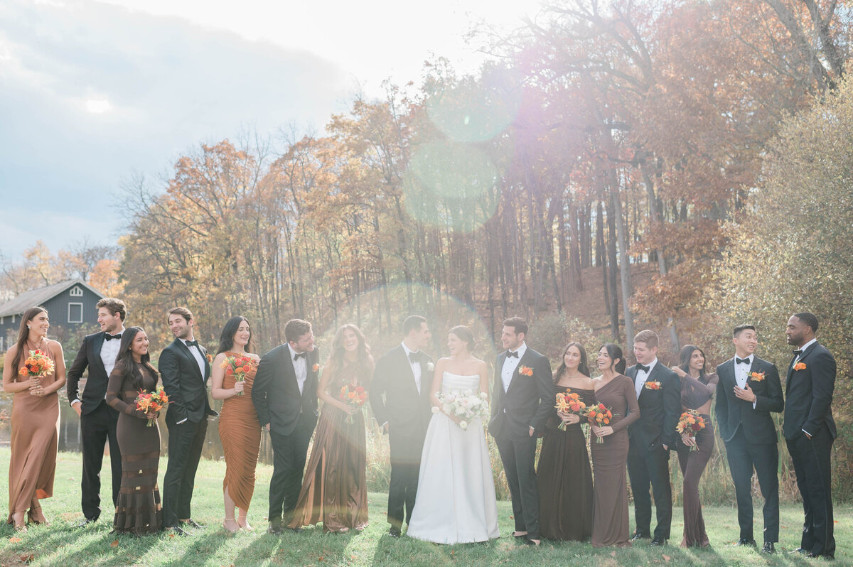  the wedding party standing in a green field with autumn foliage in the background