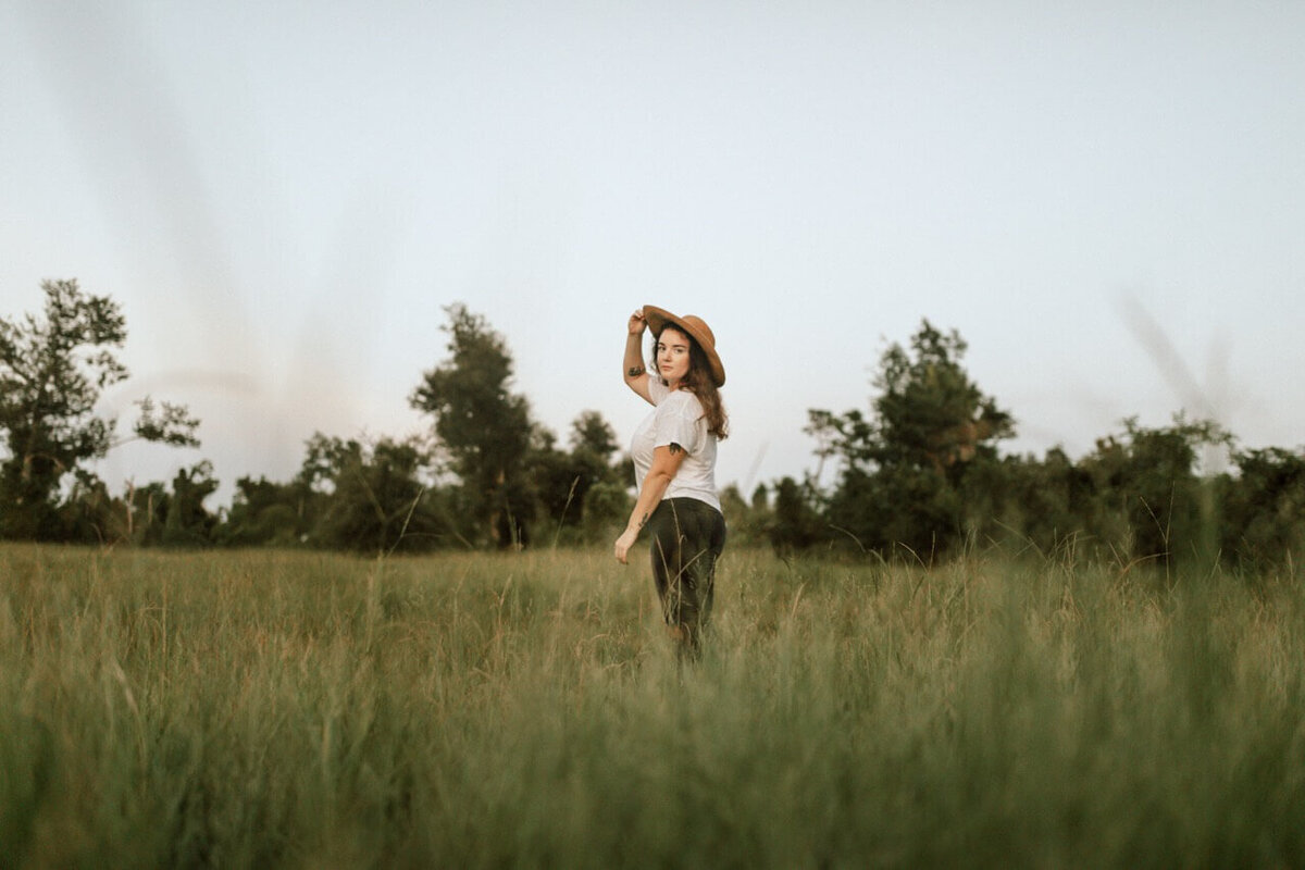woman wearing black jeans & white top posing in field with wispy grass, grabbing her hat and smiling
