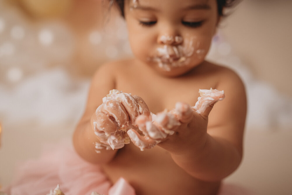 Photo shoot at Atlanta, Georgia cake smash photographer studio with one year old little girl in pink tutu smashing a cake with rainbow pastel balloon garland behind her with white fluffy clouds.