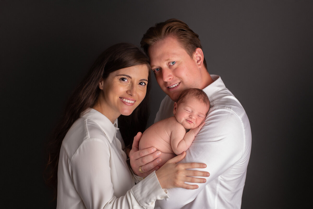 Parents facing each other with their newborn between them wearing white shirts on a dark grey background.
