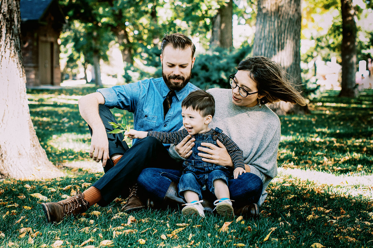 parents look down at their young son as they sit on grass at a park